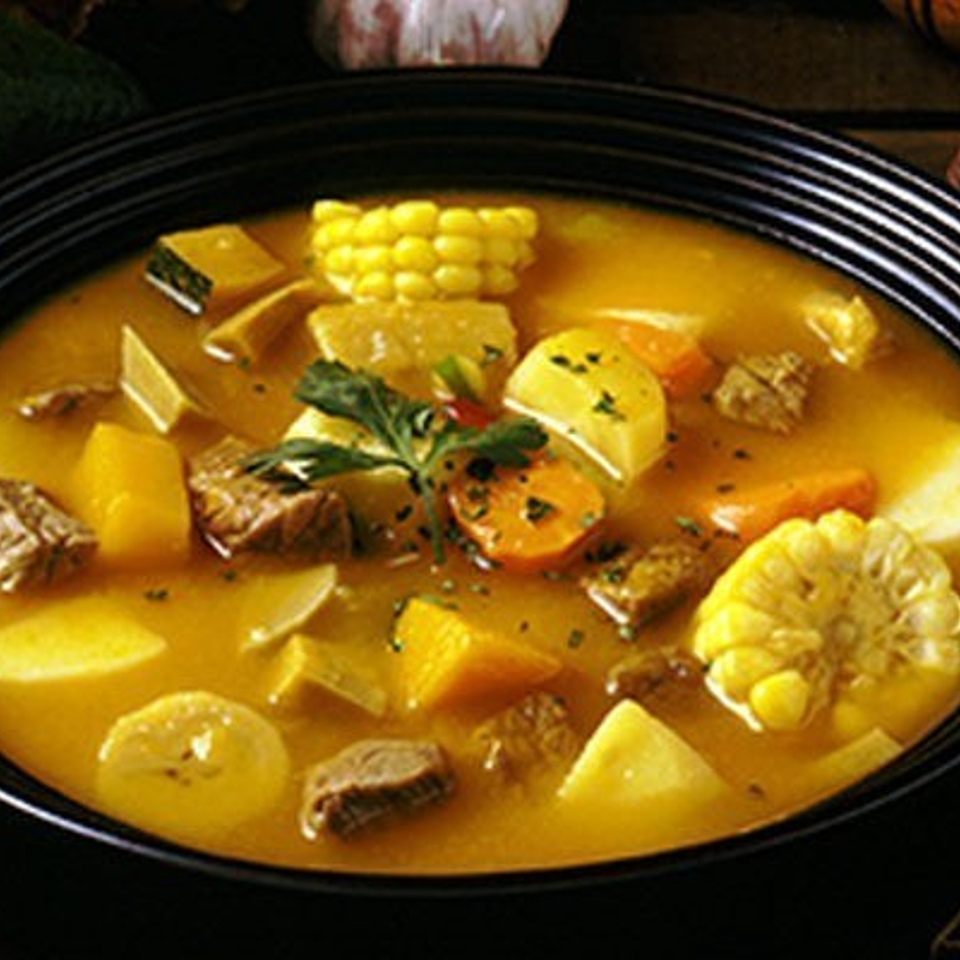 Bowl of golden vegetable and beef stew with corn, potatoes, carrots, and squash, surrounded by fresh produce and salt.
